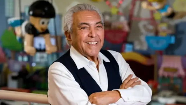 Mauricio de Sousa sorrindo, um homem idoso com óculos e cabelos brancos, em um ambiente que sugere ser sua casa ou escritório.